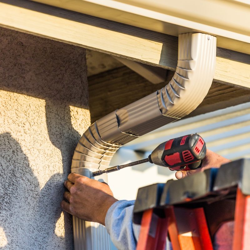 Worker Attaching Aluminum Rain Gutter and Down Spout to Fascia of House Worker Attaching Aluminum Rain Gutter and Down Spout to Fascia of House
